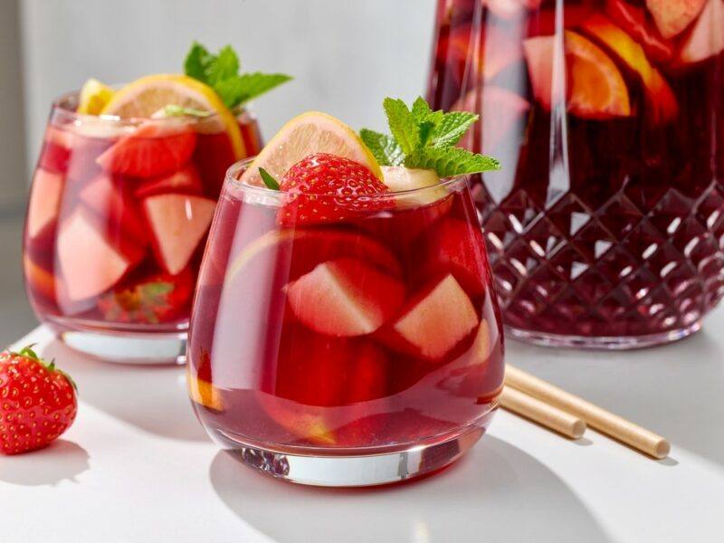 Two glasses of a trash can punch cocktail, which is filled with plenty of fresh fruit, with a jug of the punch in the background