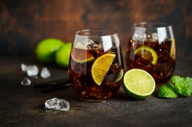 Two glasses containing whiskey and coke, with ice and lime, with more ice and limes on the table