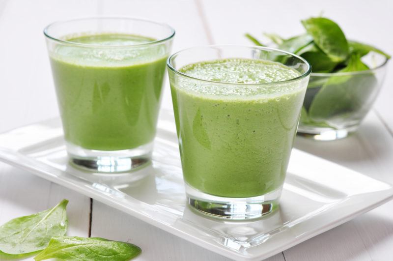 Two glasses of a green alkaline smoothie on a white plate with greens in the background