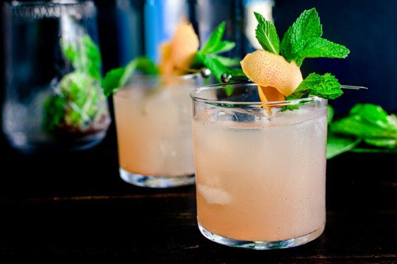 Two greyhound cocktails on a black bar with fruit and peppermint as a garnish.