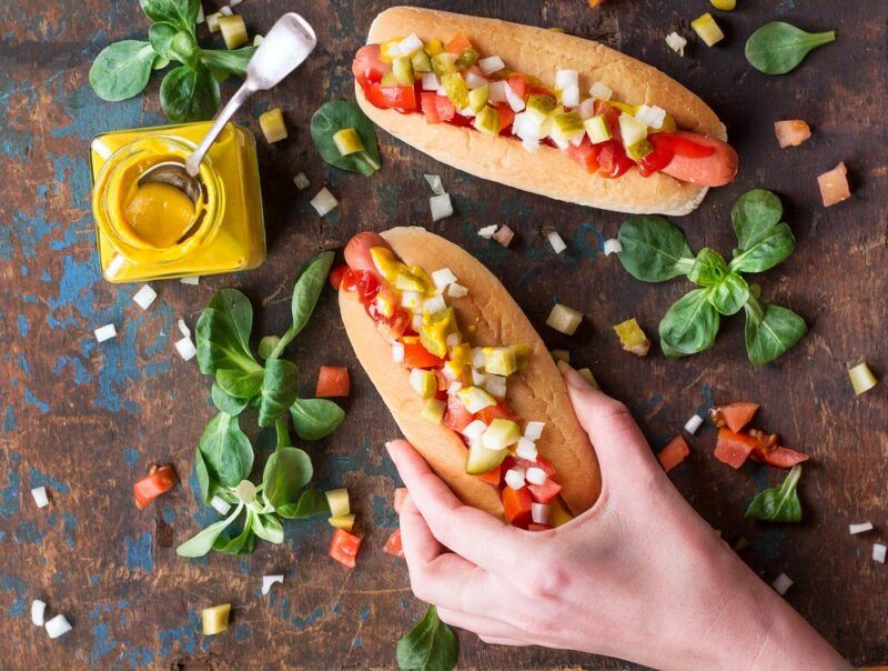 A top down view of two hot dogs. Someone is holding one of them and they both have chopped garnishes on top. There are more garnishes scattered on the table, along with a pot of mustard