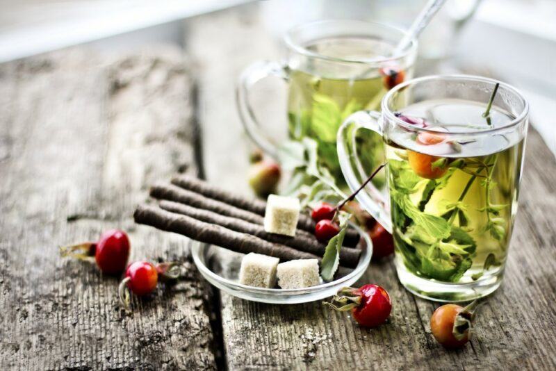 A wooden table with two mugs of herbal tea, next to a dish with sugar cubes and come cinnamon sticks, with berries scattered on the table