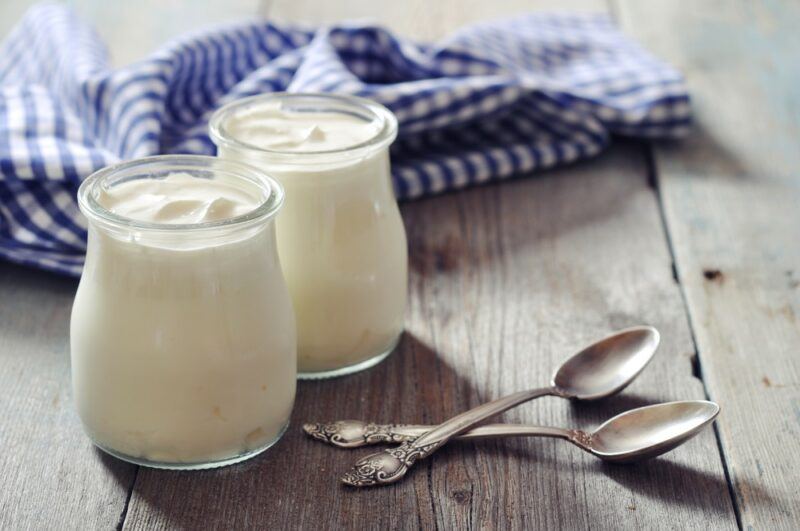 Two glass jars of Greek yogurt next to two spoons, with a blue and white cloth in the background