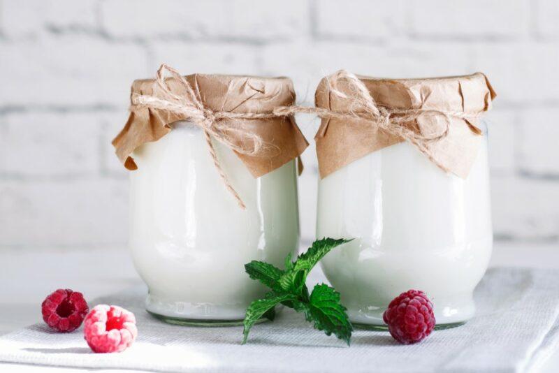 Two jars of kefir with brown paper and string at the top, in front of a white brick wall, with a few raspberries and some leaves in front