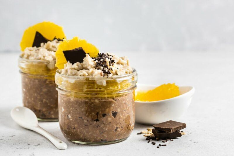 Two mason jars containing chocolate and mango overnight oats, with some chocolate and mango as garnishes and next to the jars as well