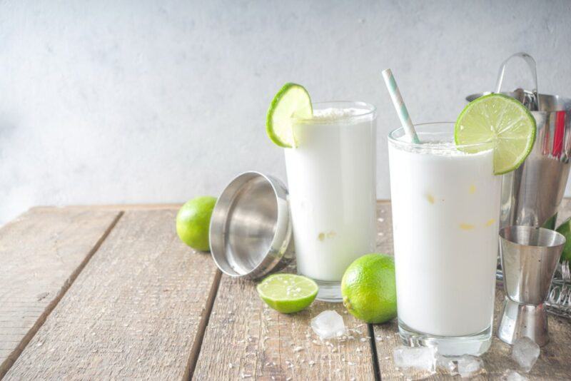 A wooden table with two tall glasses of a key lime margarita, next to many limes and a cocktail shaker