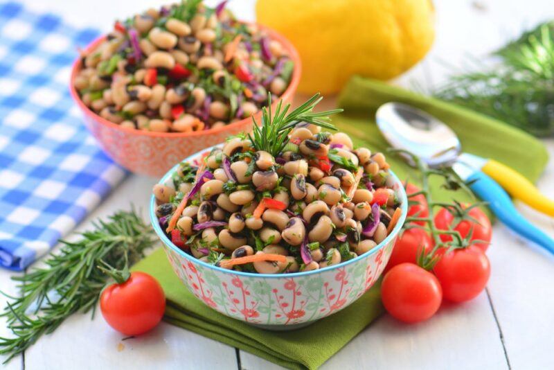 Two bowls containing a black eyed pea salad on a table with ingredients and a cloth