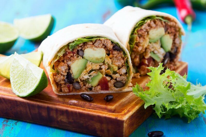 Two large burritos that include mince and avocados on a wooden board and a blue table, with lime wedges