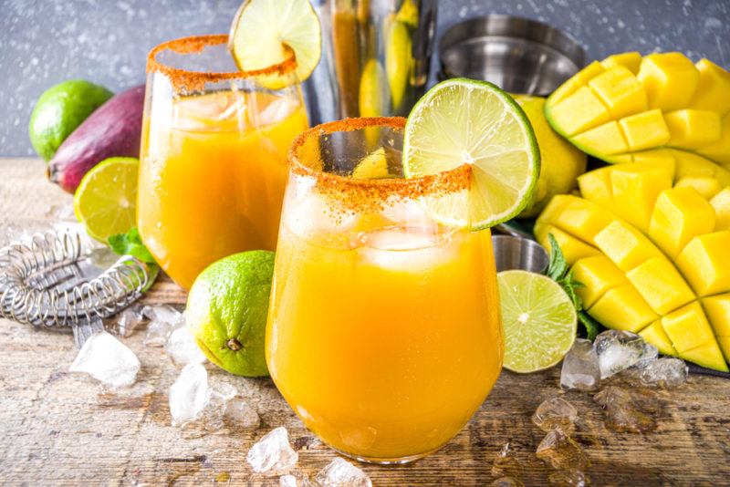 Two mango margaritas in glasses with mangos, limes, ice, and other ingredients in the background