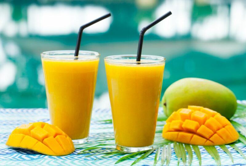 Two glasses of a mango smoothie with straw, next to two mango halves and a whole mango