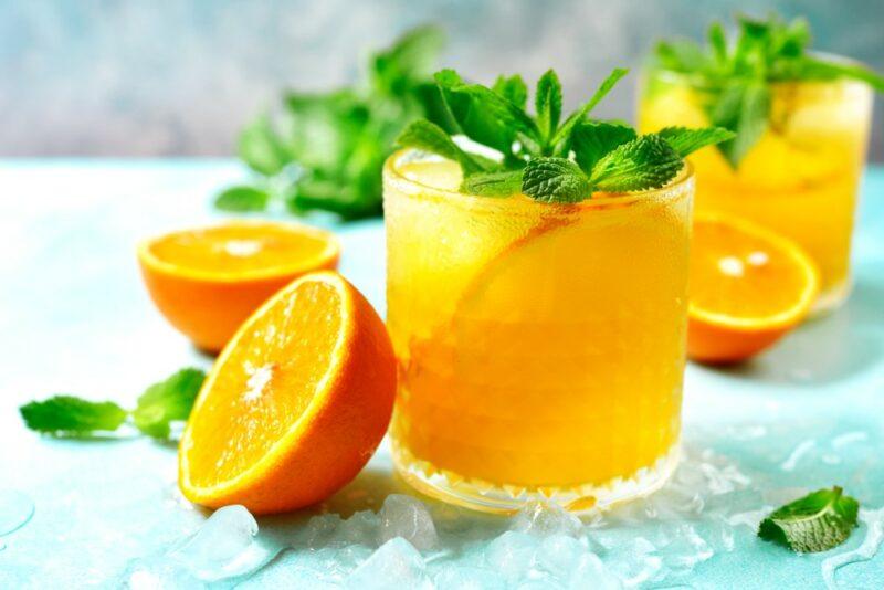 Two glasses of a manmoa cocktail on a light blue table, with oranges and peppermint leaves