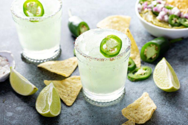 Two glasses containing margarita cocktails with a slice of jalapeno each, plus nacho chips and lime wedges on the table