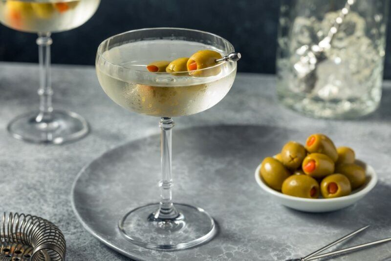 A gray plate with a martini and a bowl of olives, with another martini in the background. Both martinis are garnished with olives