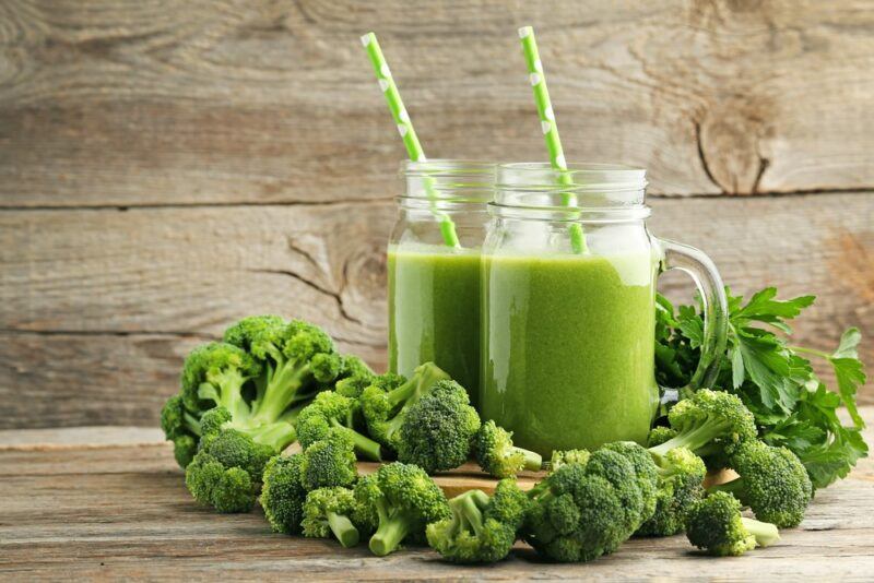 A wooden table with two mason jars of a green smoothie that uses broccoli, with straws coming out of the top