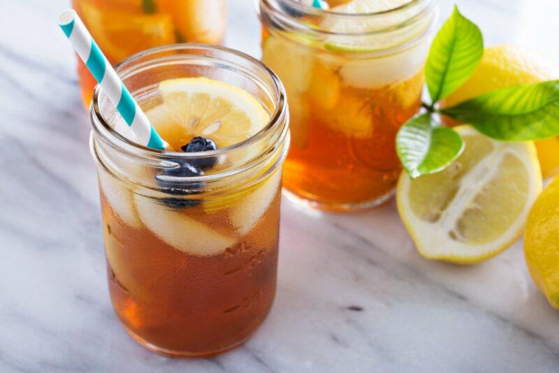 A wooden table with three mason jars containing cold brew iced tea, with ice, fruits, and a straw