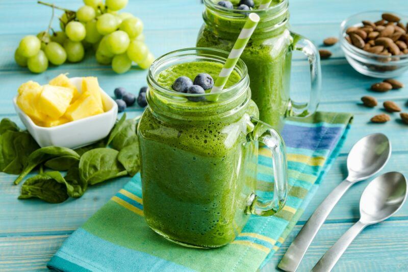 Two mason jars containing green smoothies, with pineapple pieces, grapes, almonds, blueberries, spinach, and two spoons on a table