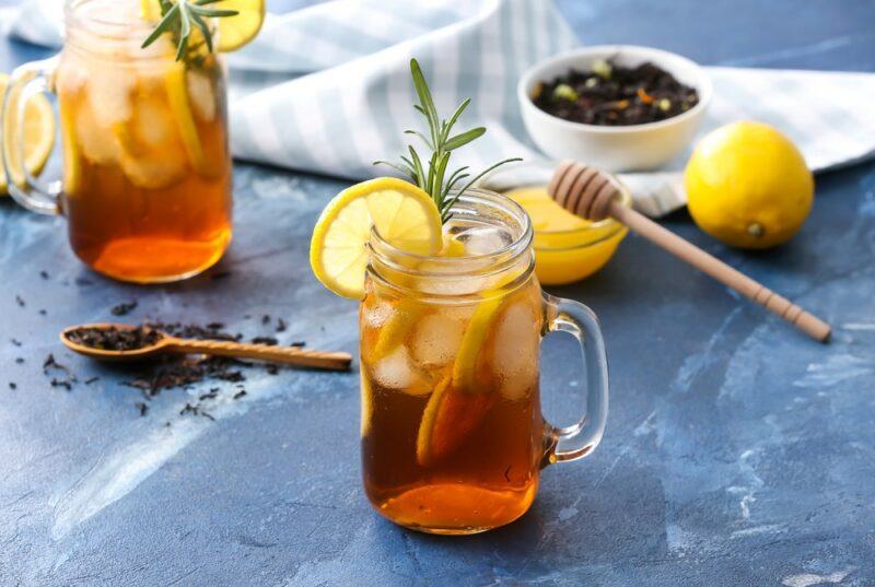 A light colored table with two mason jars containing ice, tea leaves, lemon, and honey