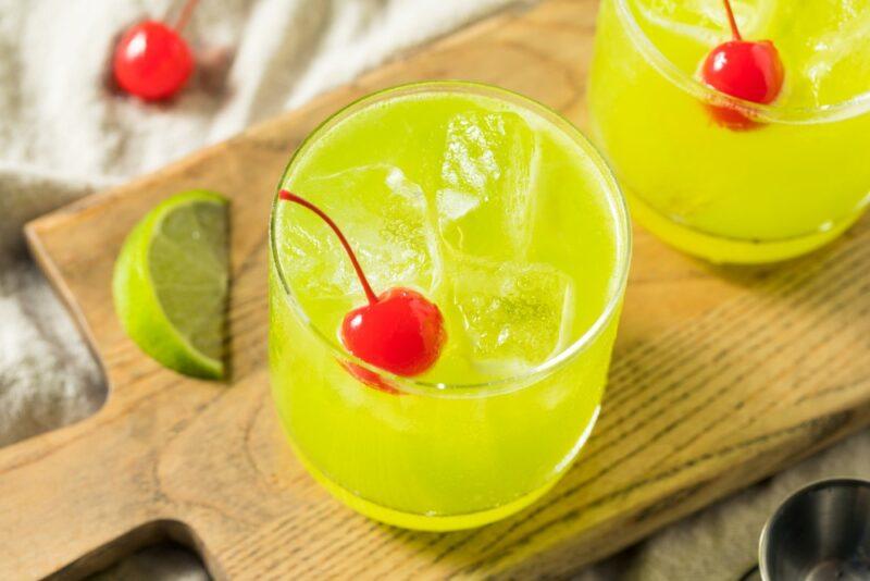 A wooden board with two vibrant melon patch cocktails, each garnished with a glace cherry