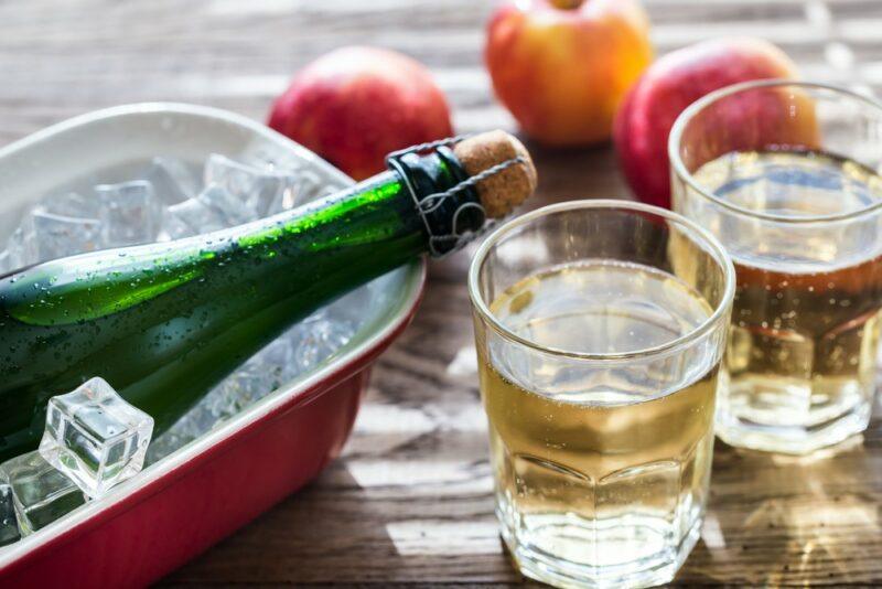 Two glasses containing a mountain cider high cocktail, in front of apples and next to a container with ice and a bottle of hard apple cider