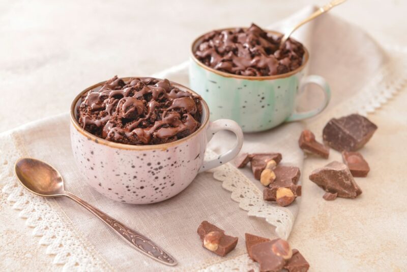 Two mugs containing chocolate cake, with chocolate chips as a garnish, next to a spoon and pieces of chocolate