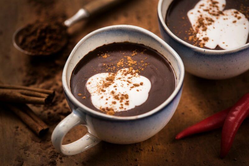 Two ceramic mugs filled with hot chocolate and topped with cream and cinnamon, on a table with cocoa powder and cinnamon