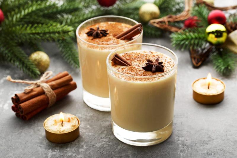 Two mugs of eggnog on a table with candles, part of a Christmas tree, and cinnamon