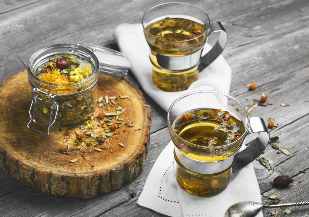 Two glass mugs of herbal tea next to a glass container of the tea on a piece of wood