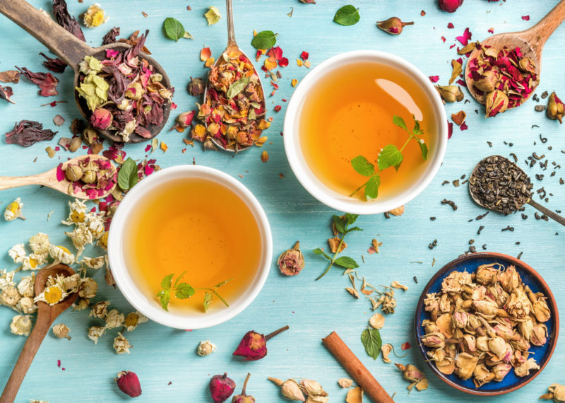 Two mugs of herbal tea next to herbs, flower petals and flower buds