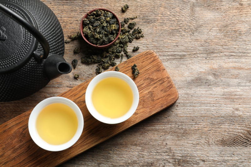 A wooden board with two white mugs filled with white tea, next to white tea leaves and a teapot