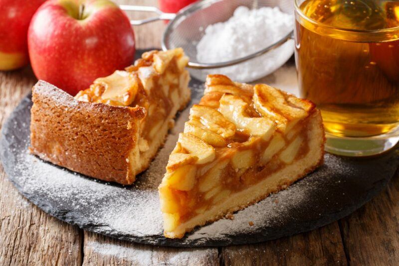 Two pieces of deep dish apple pie, next to apple juice, some apples, and a bowl of cream