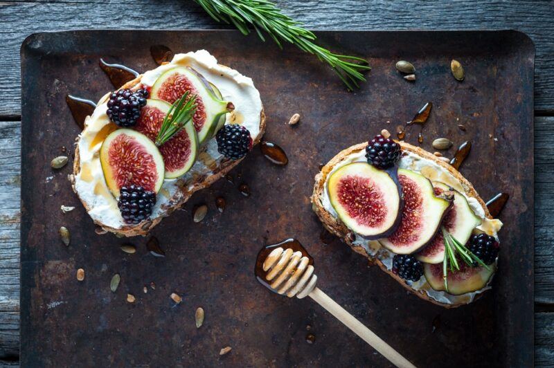 Two slices of bread with cream cheese, figs, and honey, on a black tray with a honey stirrer