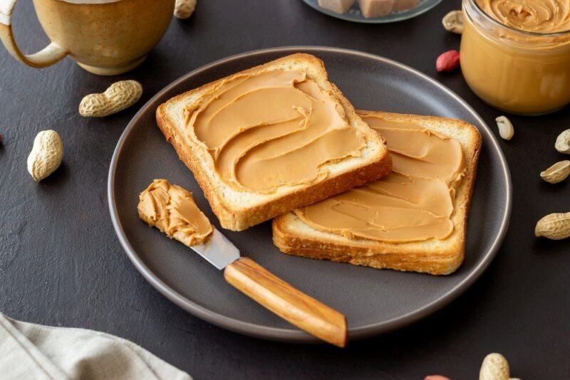 A gray plate with two pieces of toast that have been spread with peanut butter, plus a knife containing more peanut butter