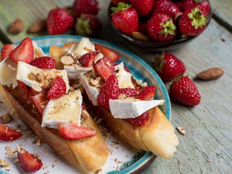 Two slices of bread with strawberries and cheese as toppings, with more strawberries on the table