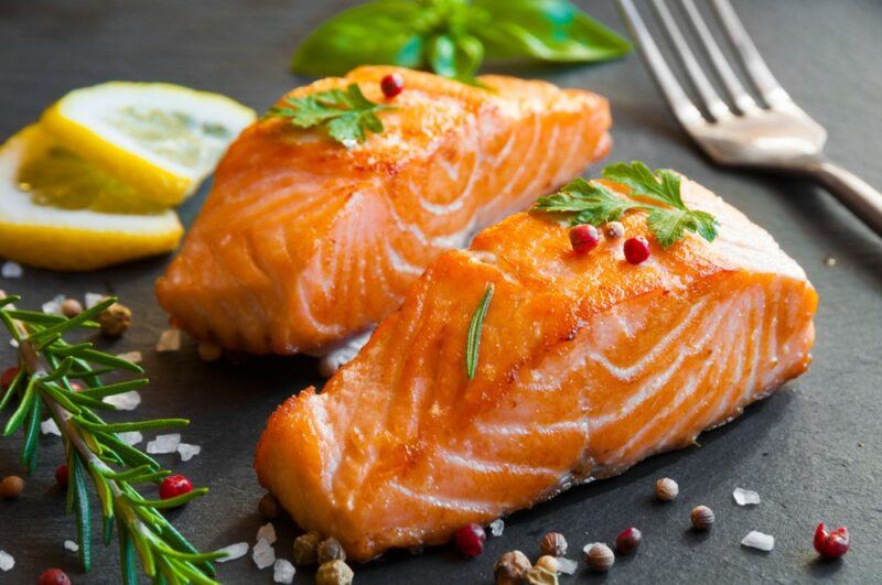 A black plate with cooked pieces of salmon, with peppercorns, rosemary, and lemon slices