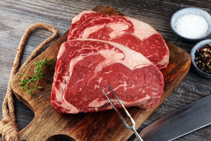 A wooden board with two rib eye steaks and a little rosemary, next to small containers of salt and pepper