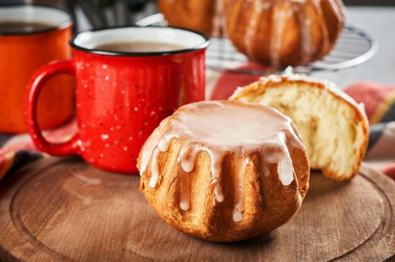 A wooden table with two pieces of rum baba cake dripping with icing, with two ceramic red coffee mugs and some more cakes in the background