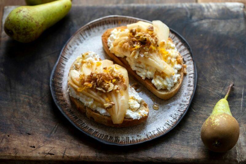 A plate with two slices of sourdough that have been topped with ricotta cheese, honey, pears, and walnuts