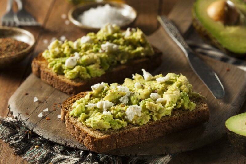 Two pieces of toast with avocado and cheese, with some salt and avocados in the background