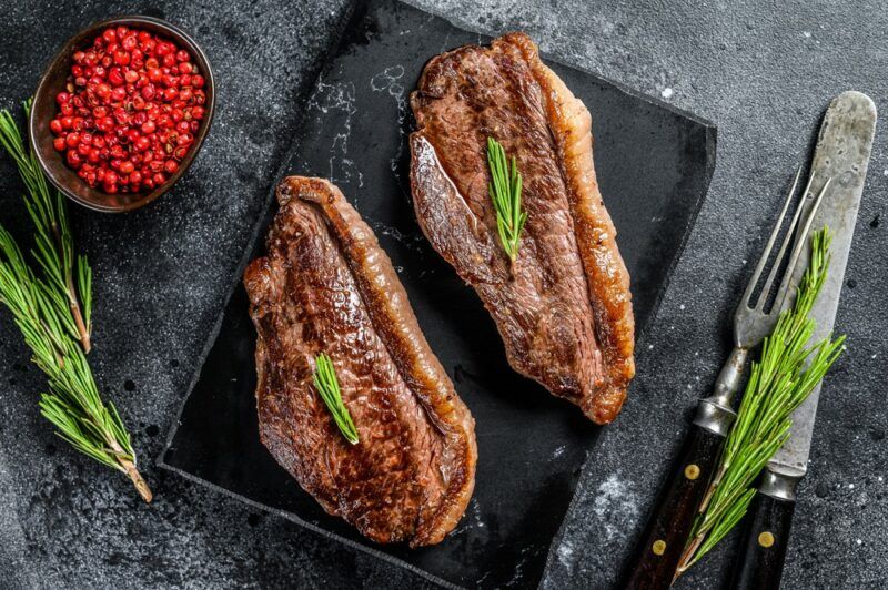 A black board with two cooked pieces of sirloin steak, next to red peppercorns, salt, and rosemary