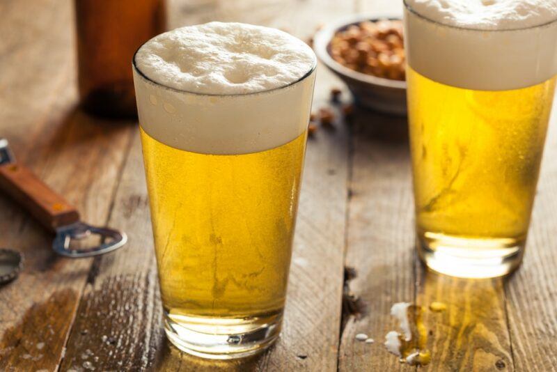 A wooden table with two large pint glasses of light beer, next to a bottle and a small dish