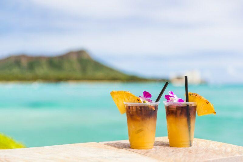 Two plastic cups containing a mai tai cocktail outside on a bench overlooking the blue sea