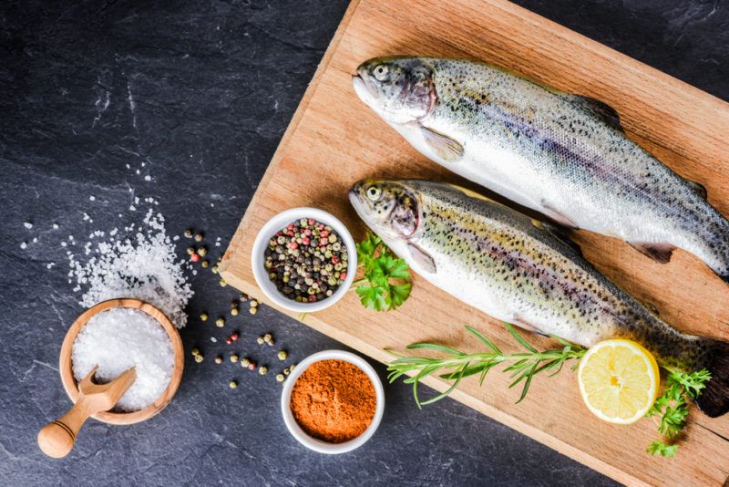 Two rainbow trout on a wooden board with small white bowls of seasoning and salt