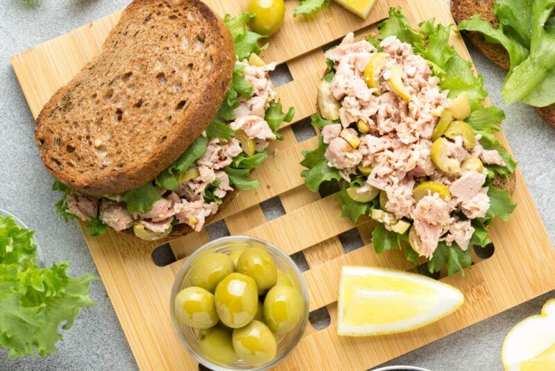 Two tuna sandwiches next to some olives on a wooden board