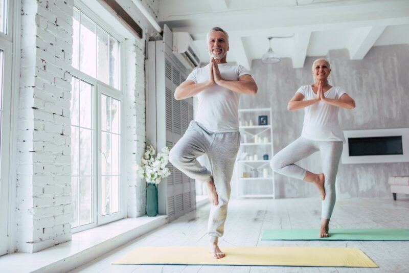 Two seniors wearing white, practicing yoga in a brightly lit studio