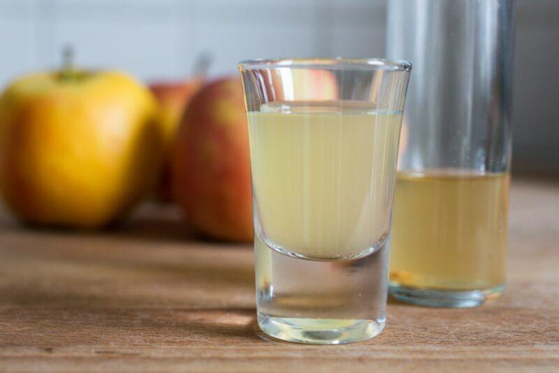 Two shot glasses, one filled with apple cider vinegar, one with the vinegar after it has been diluted. There are apples in the background