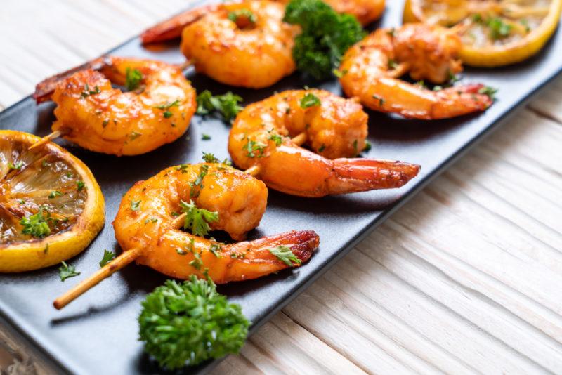 A black plate with two skewers of grilled shrimp, some parsley and a lemon slice