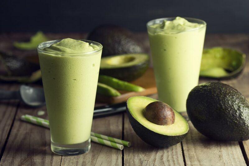 Two avocado smoothies on a wooden table with avocados