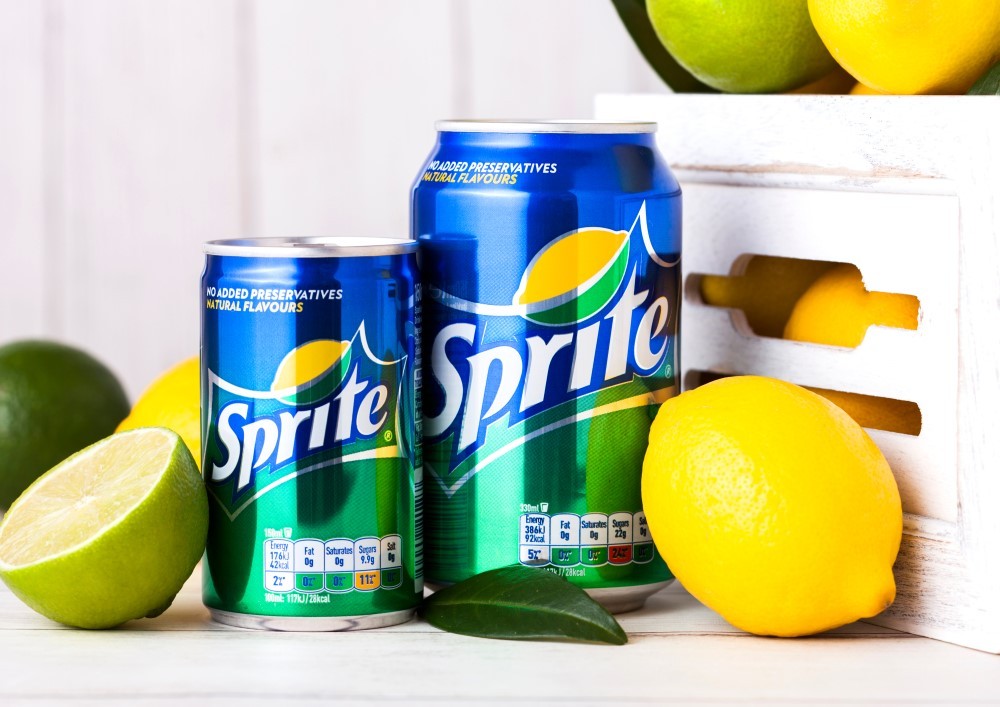 Two differently sized cans of sprite near some lemons and limes, along with a white crate