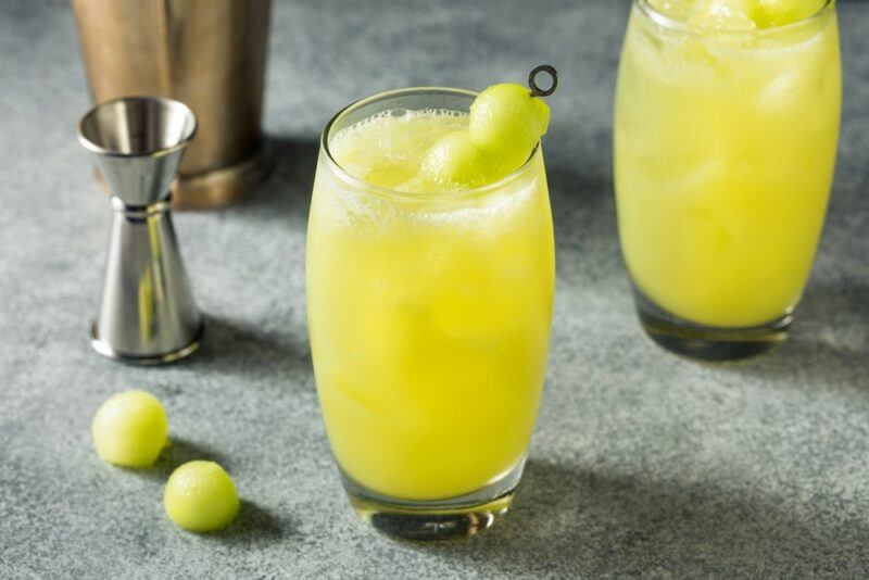 Two glasses containing a melon ball cocktail on a table with a jigger and two melon balls