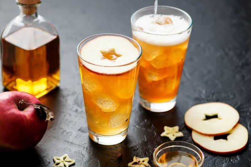 Two tall glasses of a pulp fiction cocktail with apple slices, next to a spirit and apples